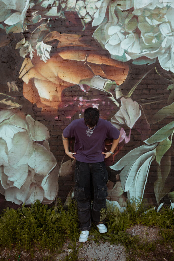 Senior guy in purple shirt poses and looks down while leaning against a graffiti floral wall with senior portraits kansas city photographer.