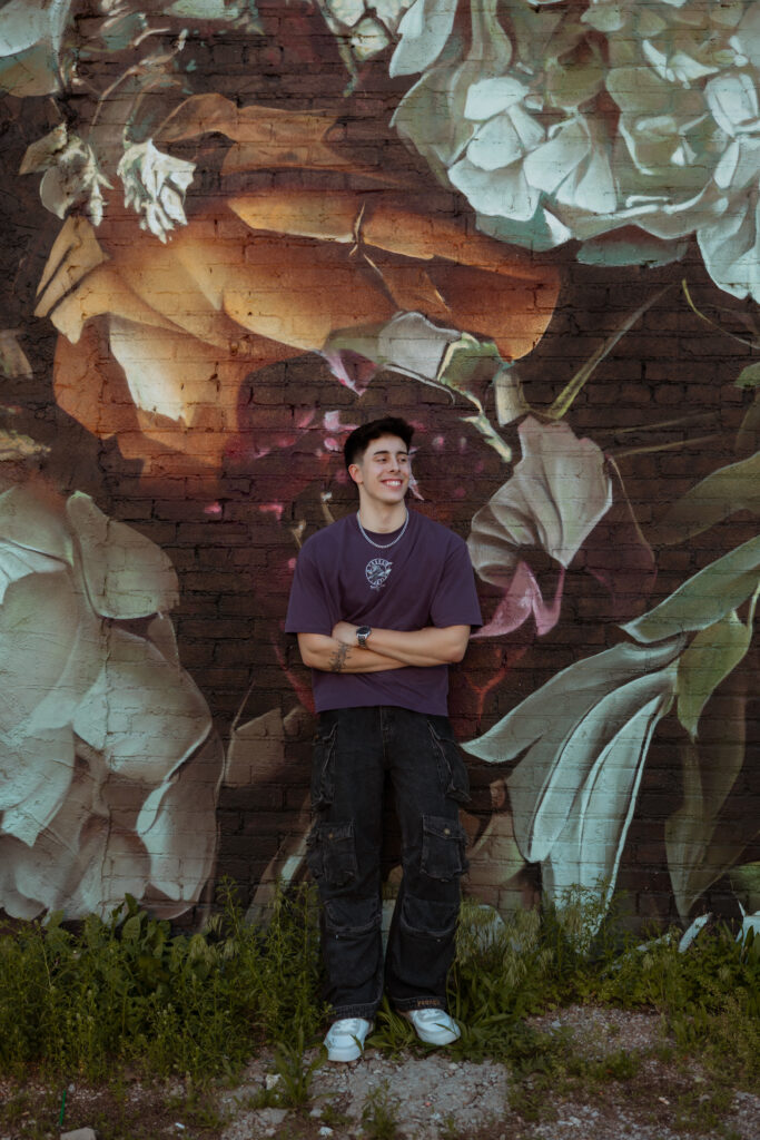 Senior guy in purple shirt poses and laughs off to the side for a shot while leaning against a graffiti floral wall with senior portraits kansas city photographer.