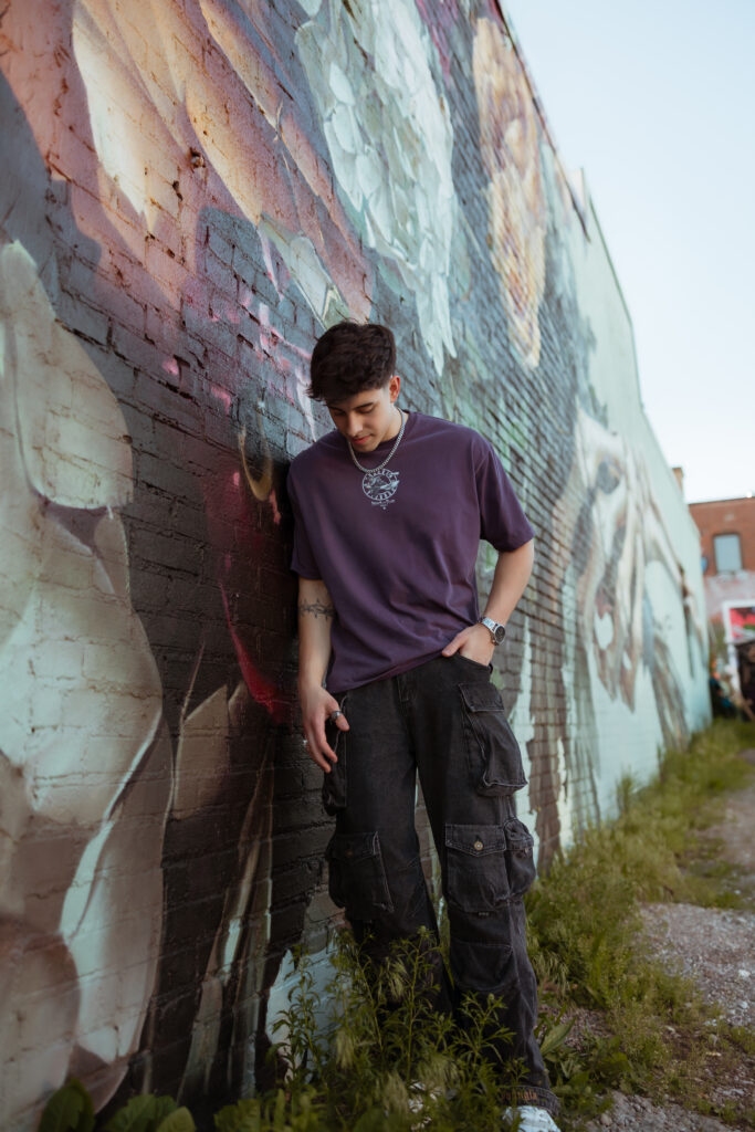 Senior guy in purple shirt leans on his shoulder against a graffiti floral wall while looking down with senior portraits kansas city photographer.