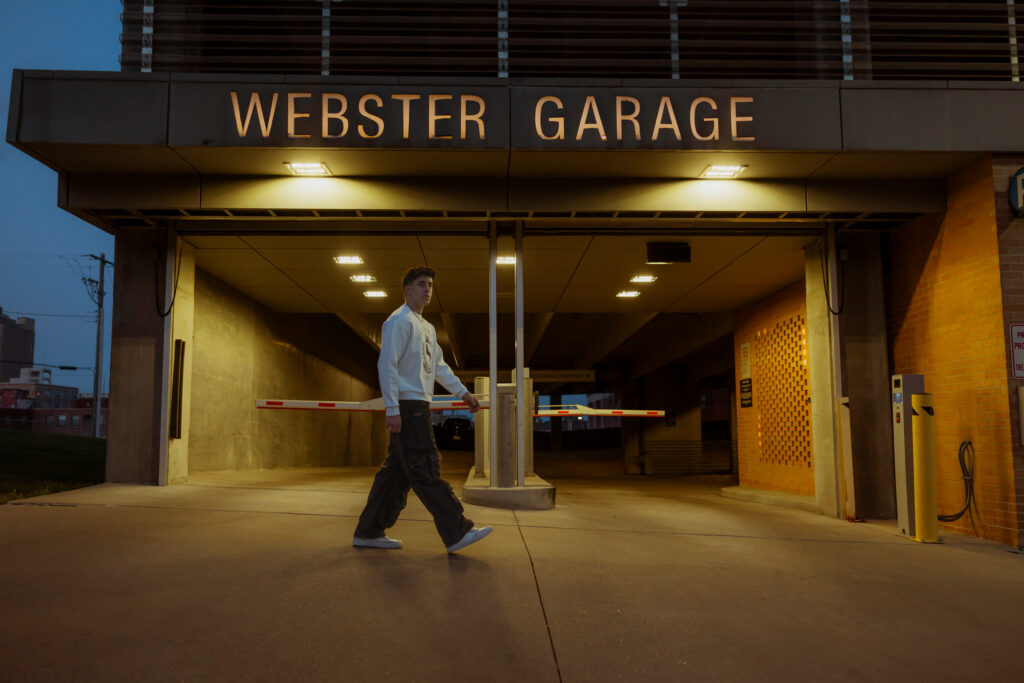 Senior portraits in Kansas City include a senior guy in white sweatshirt and black jeans walking by the opening of a parking garage in Kansas City.