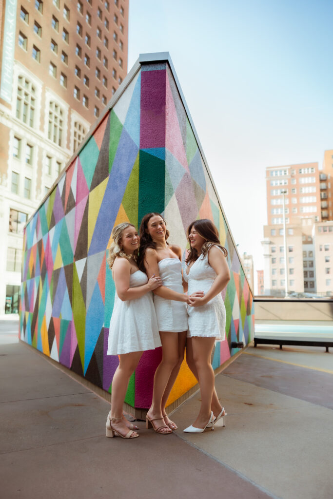 Three senior girls in white dresses laugh with each other against a colorful geometric wall with senior photography overland park ks photographers.