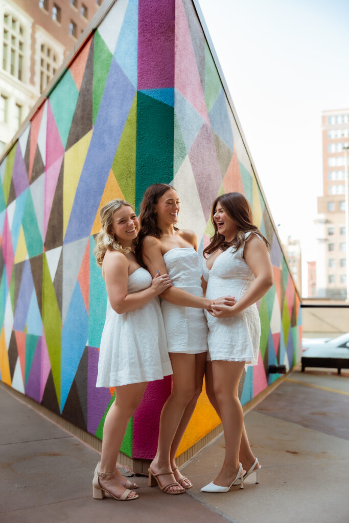 Three senior girls in white dresses laugh with each other against a colorful geometric wall with senior photography overland park ks photographers during urban senior session.