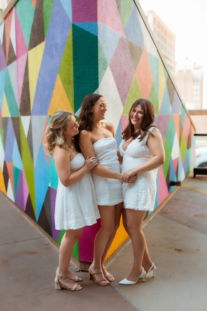 Three senior girls in white graduation dresses hold hands and laugh with each other against a colorful geometric wall with senior photography overland park ks photographers during urban senior session.