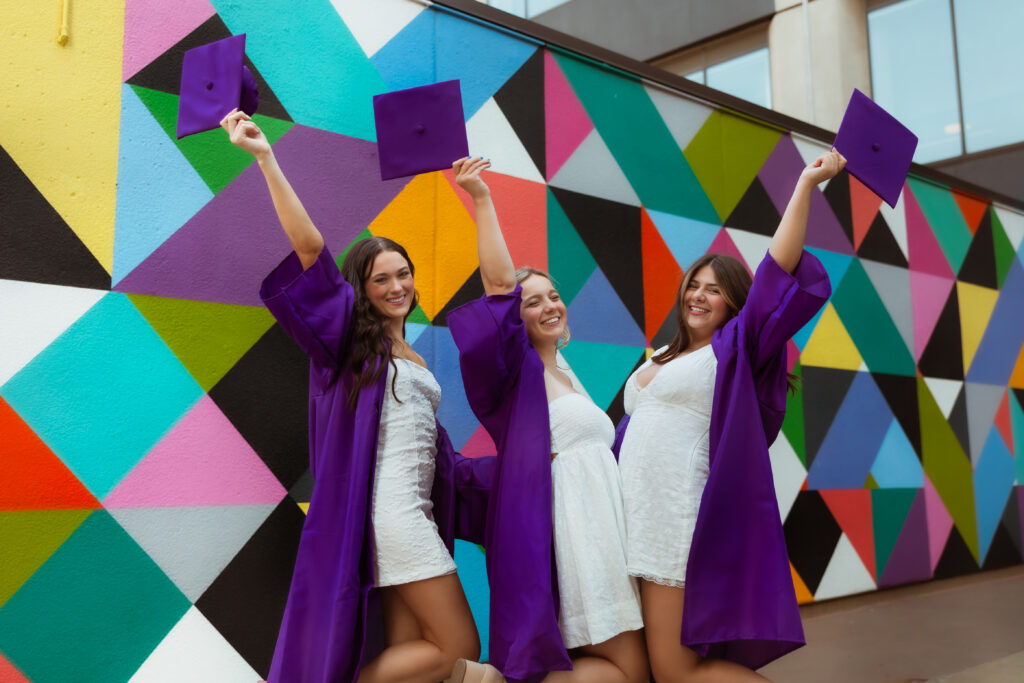 Three senior girls in their purple graduation cap and gowns and white graduation dresses hold their caps and smile against a colorful urban wall with photography overland park ks photographers during senior downtown photoshoot.