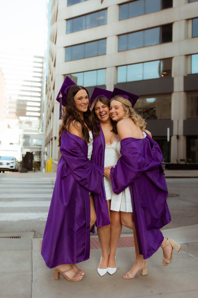Three senior girls in their purple graduation cap and gowns and white graduation dresses laugh and hug each other at crosswalk with photography overland park ks photographers during senior urban photoshoot.