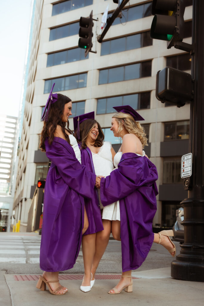 Three senior girls in their purple graduation cap and gowns and white graduation dresses laugh at each other at crosswalk with photography overland park ks photographers during senior urban photoshoot.