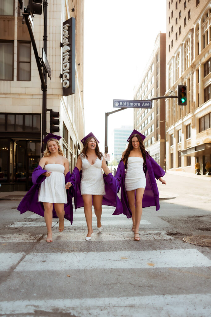 Three senior girls in their purple graduation cap and gowns and white graduation dresses laugh as they walk across a cross walk with photography overland park ks photographers.