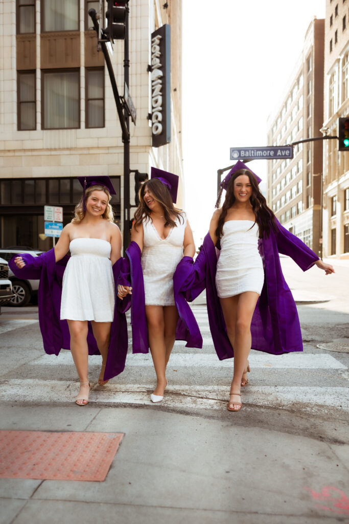 Three senior girls in their purple graduation cap and gowns and white graduation dresses laugh as they walk across a cross walk with photography overland park ks photographers during senior urban photoshoot.