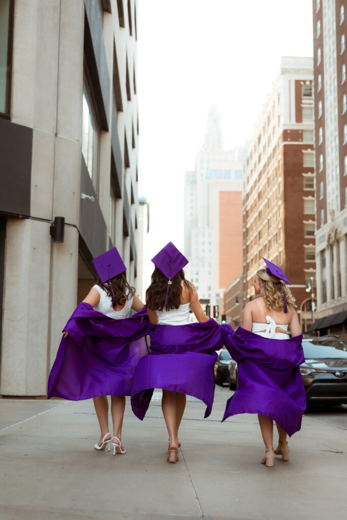 Three senior girls in their purple graduation cap and gowns and white graduation dresses walk away on sidewalk into the city distance with photography overland park ks photographers during senior downtown photoshoot.