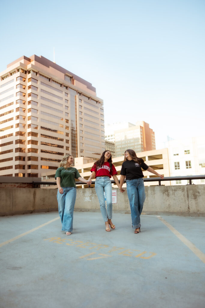 Three senior girls hold hands, wearing their college t-shirts and walk during senior session with overland park KS photographers.