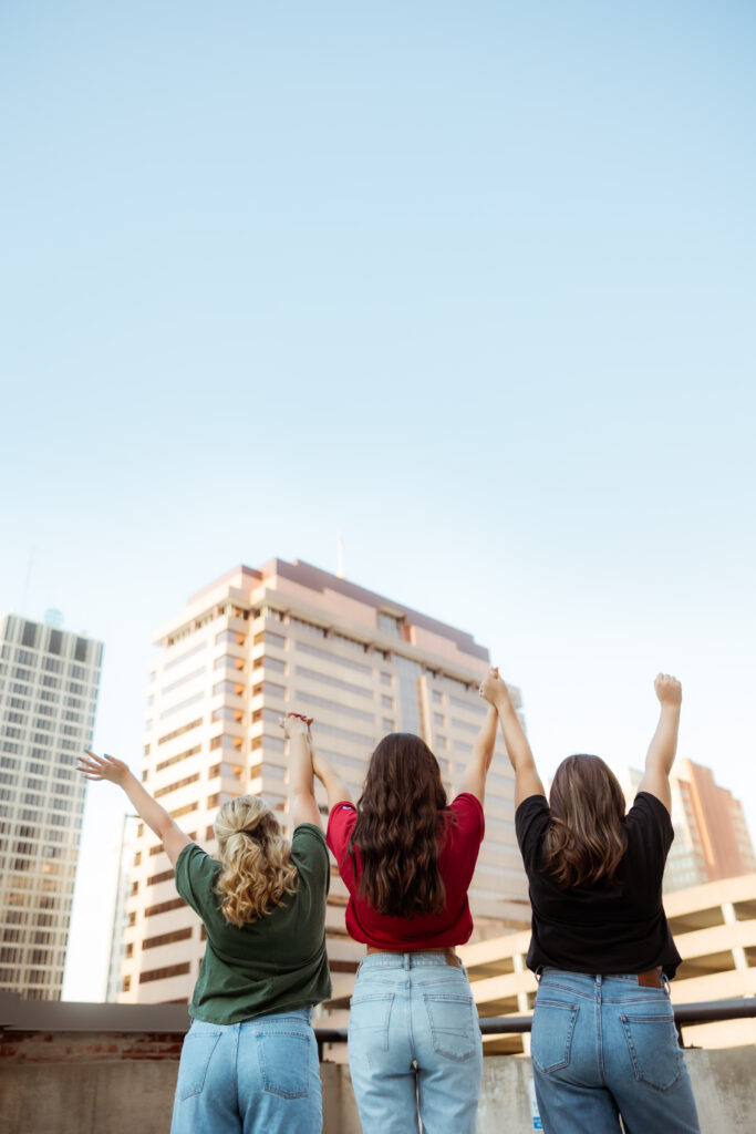 Three senior girls hold hands, wearing their college t-shirts and face away at the skyline during senior session with overland park KS photographers.