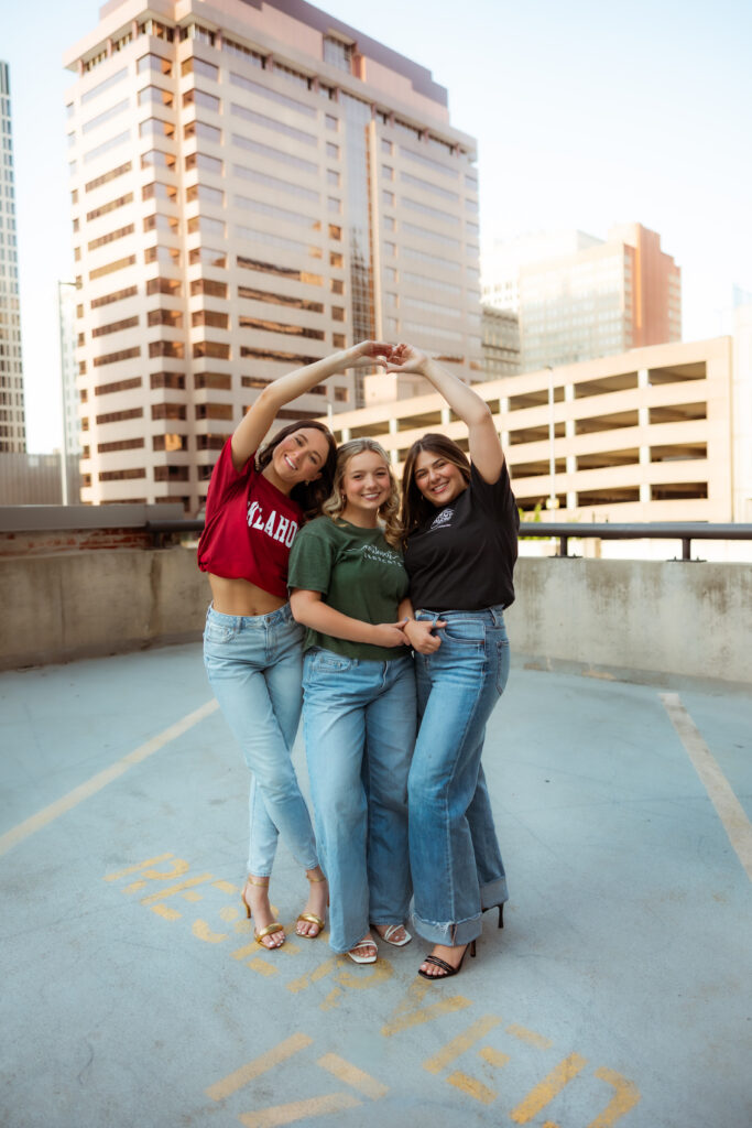 Three senior girls wearing their college t-shirts have their arms around each other and make a heart above their heads during senior session with overland park KS photographers.