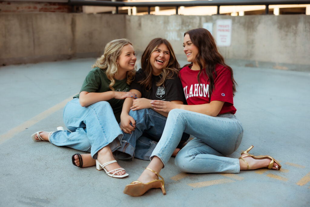 Three senior girls in college t-shirts sit on parking garage floor while they laugh at each other with overland park KS photographers.
