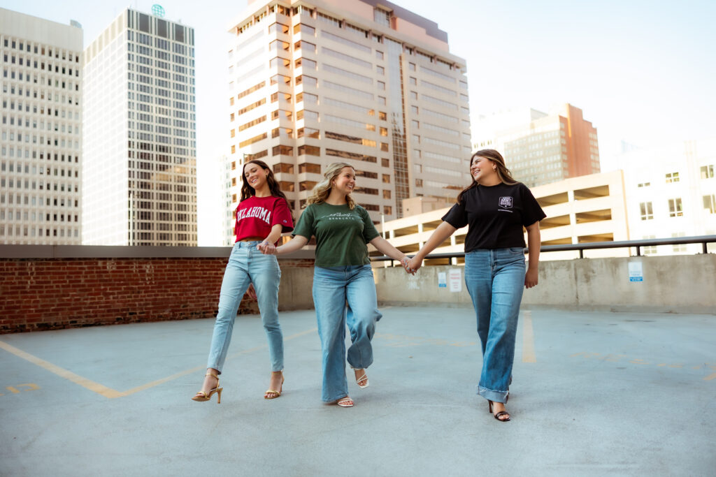 Three senior girls wearing college tees and jeans give hold hands and walk together on parking garage during senior session with photography overland park ks photographers.