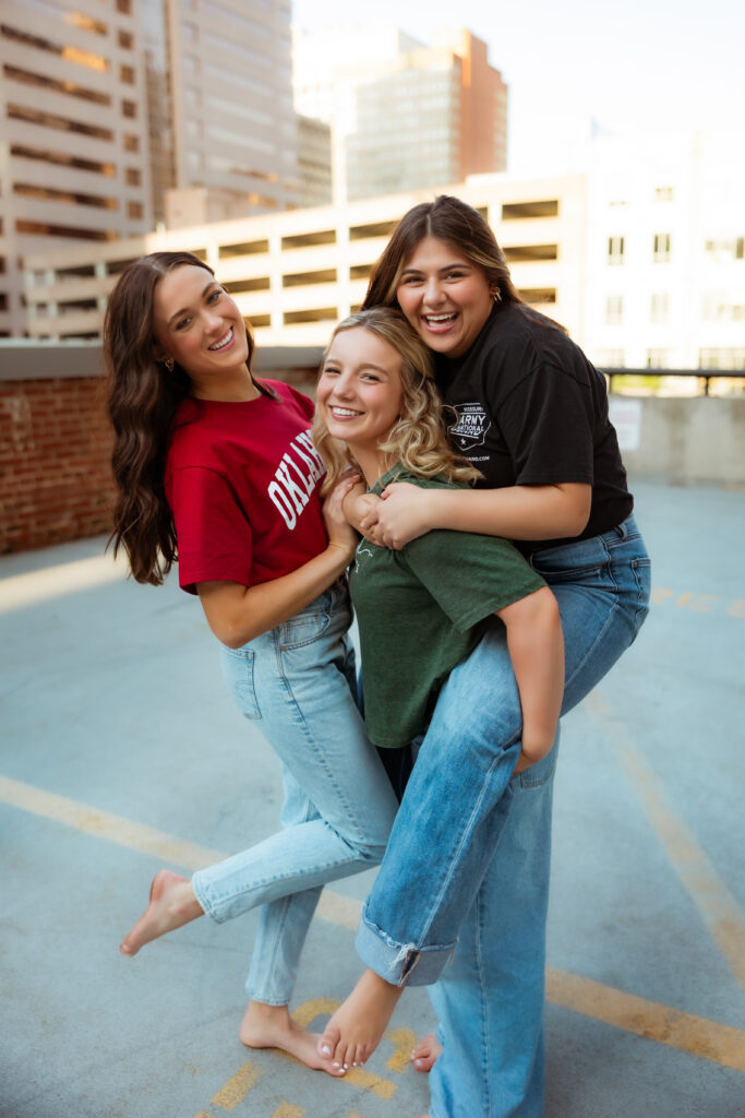 Three senior girls wearing college and military shirts and jeans give piggy back rides and laugh on parking garage during senior session with photography overland park ks photographers.