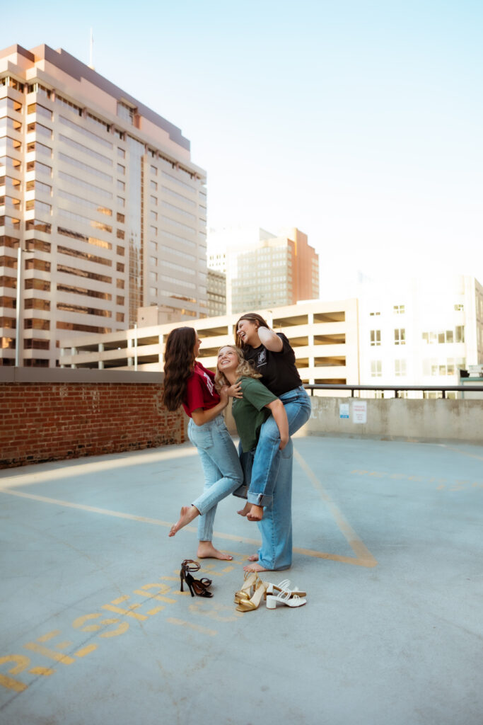 Three senior girls wearing college tees and jeans give piggy back rides and laugh on parking garage next to their high heel shoes during senior session with photography overland park ks photographers.