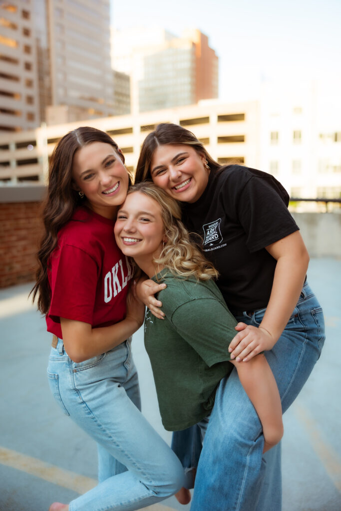 Three senior girls wearing college tees and jeans give piggy back rides and laugh on parking garage during senior session with photography overland park ks photographers.