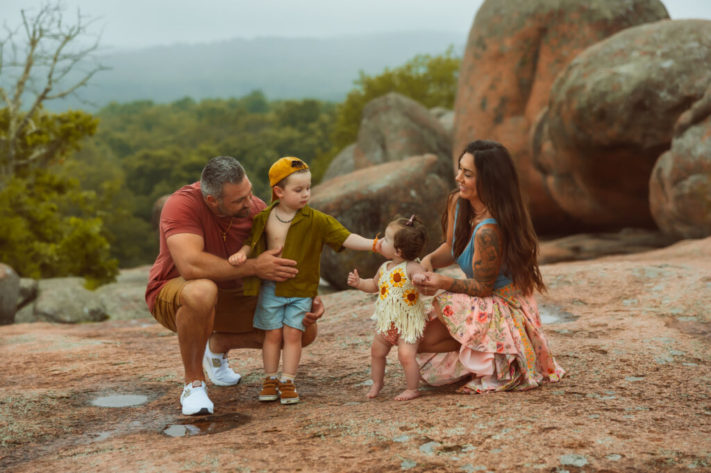 Elephant Rocks State Park Family Photographers capture dad squatting with his son and mom squatting with her daughter while the little boy puts his hand on his little sisters cheek during a sweet moment.