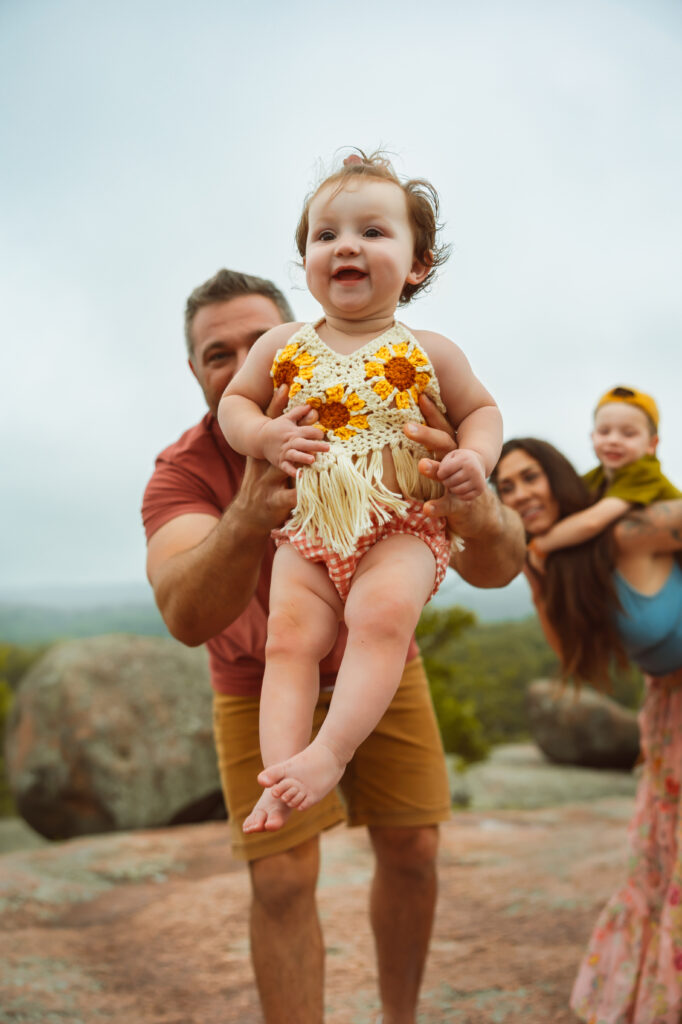 Elephant Rocks State Park Family Photographers take a photo of dad swinging his little girl towards the camera as she smiles and mom and brother are in the background.