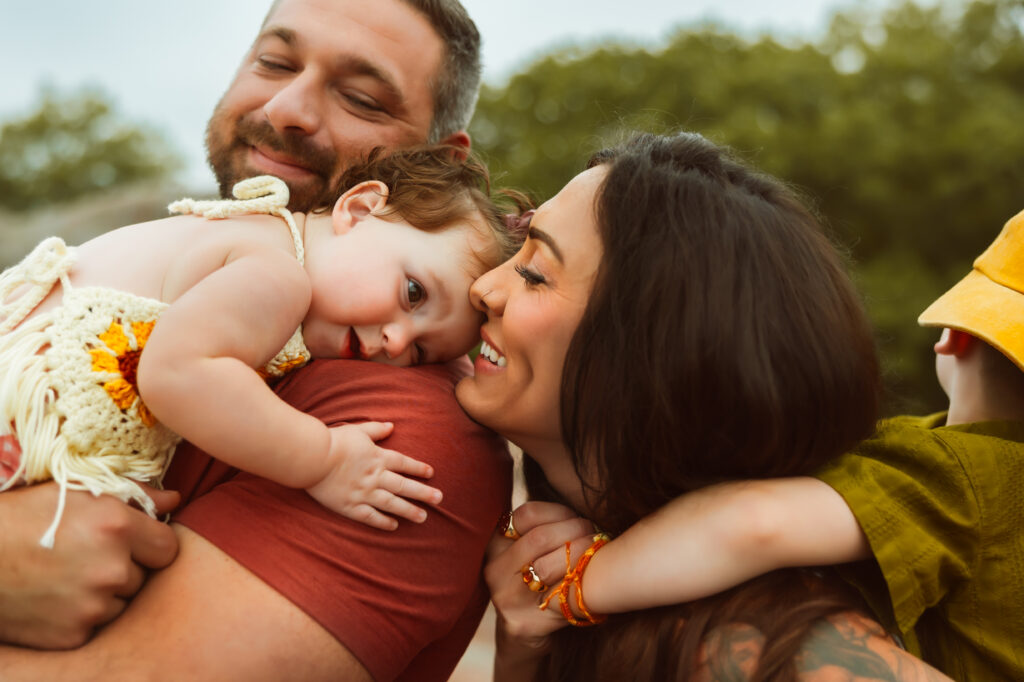 Dad holds his little girl on his shoulder as mom giving son a piggy back ride smiles and kisses daughter with Elephant Rocks State Park Family Photographers.