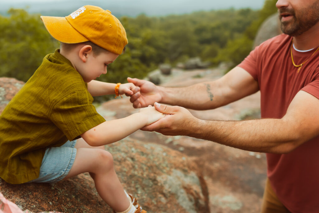 Dad hold hands with his son as his son in a green shirt and jean shorts and yellow ball cap jumps off rocks with Elephant Rocks State Park Family Photographers.