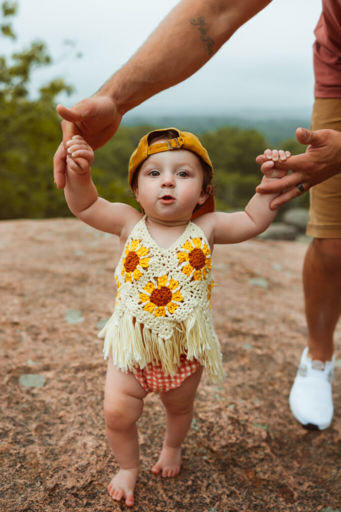 Little girl in a knit sunflower top and checkered bottoms wears her big brothers yellow hat backwards as she walks holding dad's fingers during family photos with Elephant Rocks State Park Family Photographers.